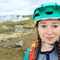 Photograph of Aoife Glass wearing a turquoise helmet and standing in front of a fumerole in Iceland